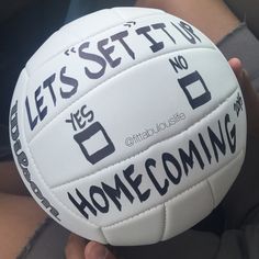 a person holding a white volleyball with words written on the side and black lettering that reads let's set up for home coming