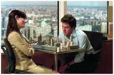 two people sitting at a table playing chess in front of a window with the city skyline behind them