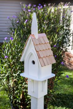 a white birdhouse with a brown roof in front of some bushes and purple flowers