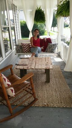 a woman sitting on a porch with a wooden table and rocking chair in front of her