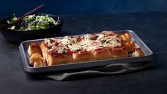 a square pan filled with food next to a bowl of salad on a black table