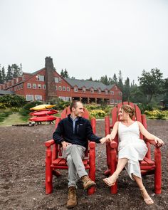 a man and woman sitting on red chairs in front of a large house with trees