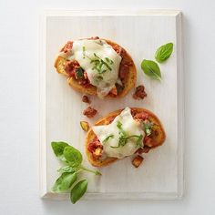 two pieces of bread with meat and cheese on them sitting on a cutting board next to basil leaves