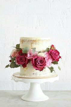 a white cake with pink and red flowers on top is sitting on a table in front of a white brick wall