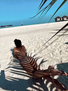 a woman laying on top of a sandy beach under a palm tree next to the ocean