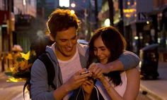 a man and woman standing next to each other on the street looking at their cell phones