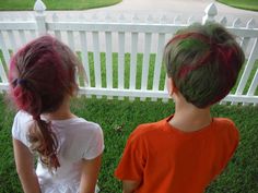 two young children with dyed hair sitting on the grass in front of a white picket fence