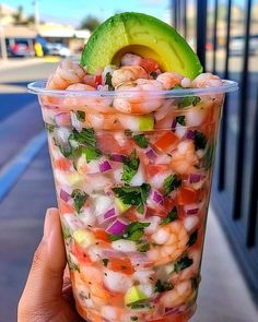 a person holding up a plastic cup filled with shrimp and veggies, topped with an avocado