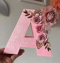 a hand holding up a pink letter decorated with flowers and paper roses in front of a white wall