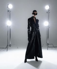 a woman standing in front of three lights wearing a long black coat and matching sunglasses