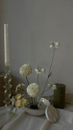 white flowers in a bowl on a table next to a candle and some candlesticks