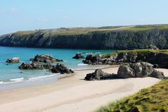 a sandy beach next to the ocean with rocks in the water and green grass on both sides