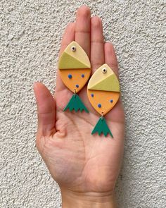 a hand holding two wooden earrings with green and yellow shapes on each earring, in front of a white wall