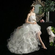 a woman in a white dress sitting next to a mirror and vase with flowers on it