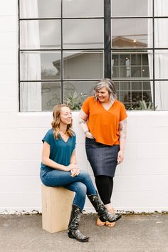 two women sitting next to each other in front of a white brick wall and windows