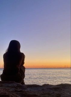 a woman sitting on top of a rock next to the ocean at sunset or dawn