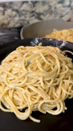 a black plate topped with pasta next to a bowl of sauce and a silver spoon