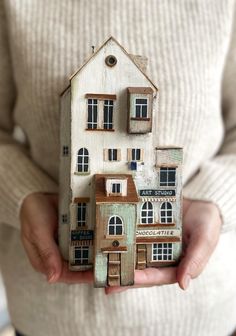 a person holding a small wooden model of a house with windows and shutters on the front