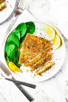 a white plate topped with slices of chicken next to spinach and lemon wedges