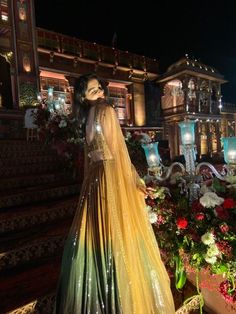 a woman in a long dress standing on some steps at night with flowers and lights behind her