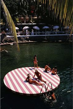 several people are in the water on a pink and white striped floating platform that is surrounded by palm trees