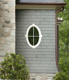 a round window on the side of a gray brick house with green trees in the background