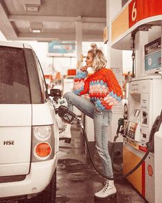 a woman is pumping gas into her car at a gas station while wearing an orange and blue sweater