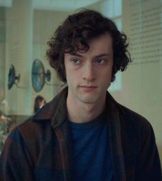 a young man with curly hair wearing a blue shirt and brown jacket looking at the camera