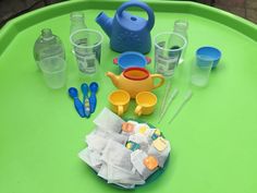 a green table topped with plastic cups and containers filled with water, teapots and utensils