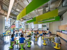 children are sitting at tables in an open area with green and yellow accents on the ceiling