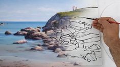 a man is drawing on a piece of paper near the ocean with his hand and pencil