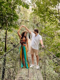 a man and woman walking across a suspension bridge in the jungle with trees around them