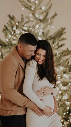 a pregnant couple standing in front of a christmas tree with their arms around each other