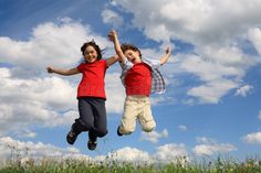 two young children jumping in the air on a sunny day with blue sky and clouds behind them