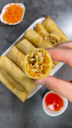 a hand holding up a vegetable spring rolls with dipping sauces on the table behind it