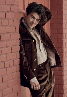 a man leaning against a brick wall with his hand on his head, wearing a brown suit and tie