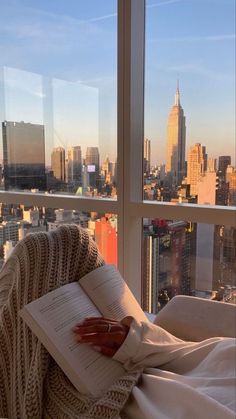 an open book sitting on top of a bed in front of a window overlooking a city