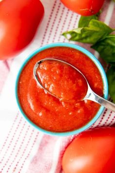 a spoon in a blue bowl filled with tomato sauce next to tomatoes and basil leaves