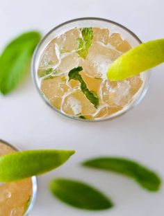 two glasses filled with ice and limes on top of a white table next to green leaves