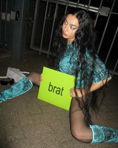 a woman sitting on the ground with her legs crossed holding a sign that says brat