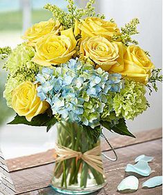 a vase filled with yellow roses and blue hydrangeas on top of a wooden table