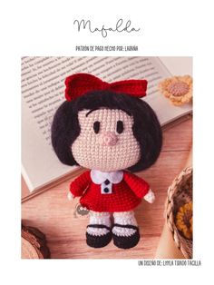 a crocheted doll with a red dress and black hair sitting next to an open book