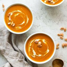 three bowls filled with carrot soup on top of a white counter next to two spoons