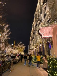 people are walking down the sidewalk in front of buildings with christmas lights on them and trees all around