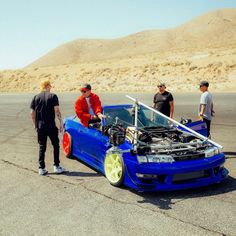 three men standing next to a blue car with the hood up and an engine on it