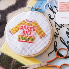 a pair of scissors sitting on top of a table next to a cross stitch shirt