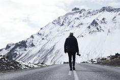 a man walking down the middle of an empty road in front of snow covered mountains