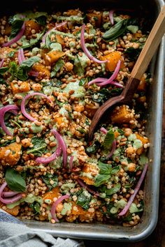 a pan filled with salad and vegetables on top of a wooden table next to a spoon