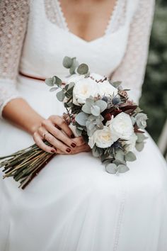 a woman holding a bouquet of white flowers and greenery in her hands, wearing a wedding dress