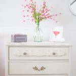 a white dresser with pink flowers in a vase on top and a clock behind it